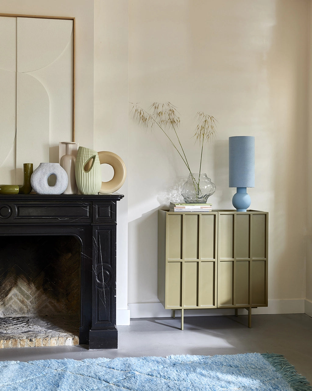 pastel blue table lamp with ball foot and tall linen shade on a olive green colored small cabinet with glass vase and flower stems