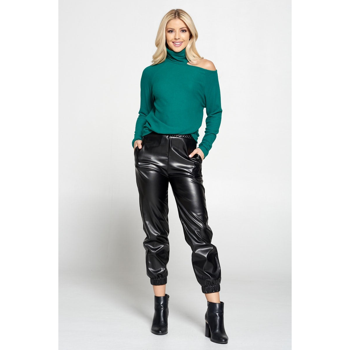 Blonde woman with long hair wearing faux black leather pants with a green cold shoulder turtleneck sweater 