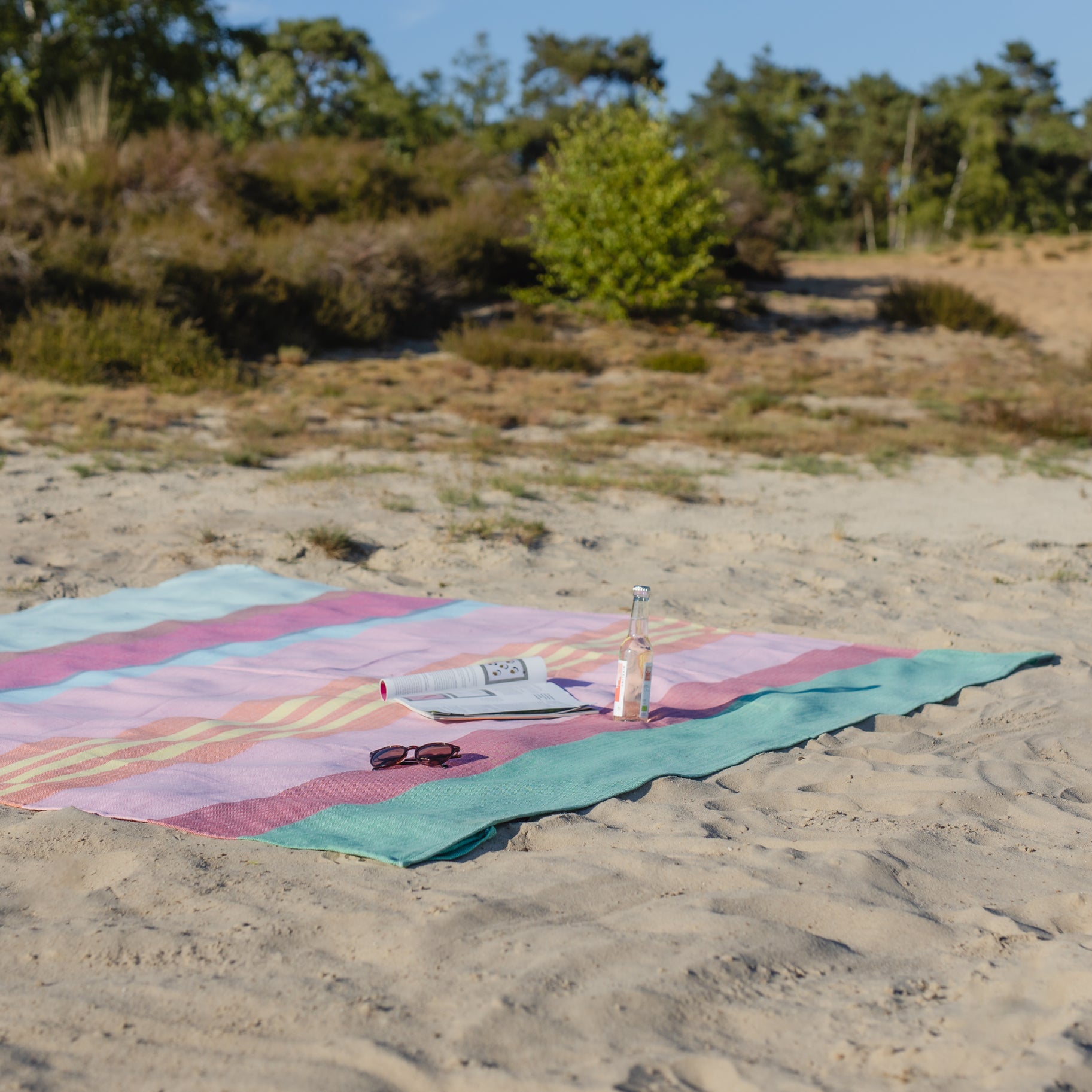 super large, striped blue, green, pink, yellow orange beach blanket on a sandy beach with a magazine and a bottle of soda