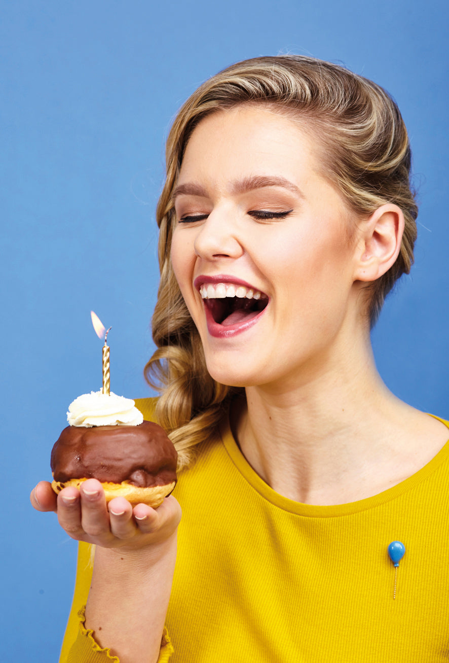 woman celebrating with cake and candle and a blue balloon pin on her yellow shirt