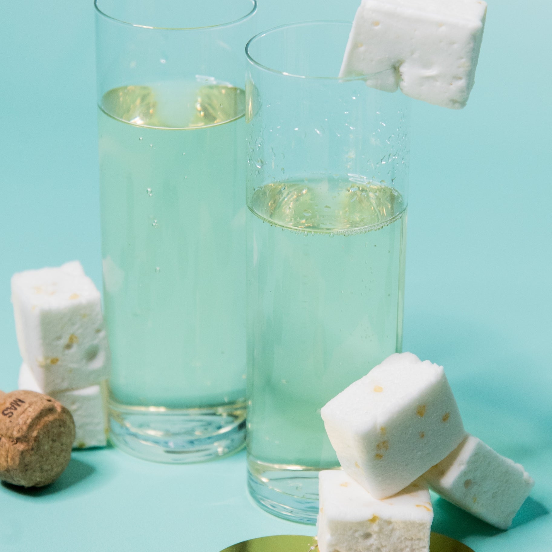 two longdrink glasses filled with champagne and marshmallow cubes against a light blue background